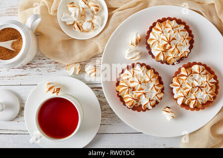 tasty meringue mini tarts with browned meringue peaks served with tea, mini marshmallows and brown organic sugar on wooden table with light cloth, vie Stock Photo