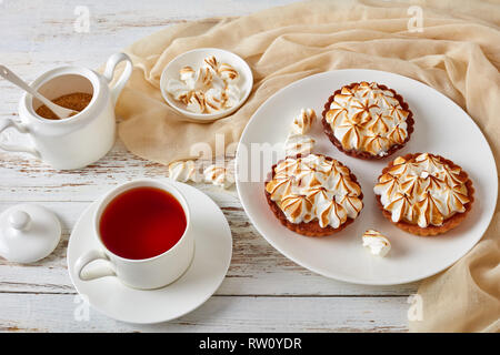 close-up of meringue mini tarts with browned meringue peaks served with tea, mini marshmallows and brown organic sugar on wooden table with light beig Stock Photo