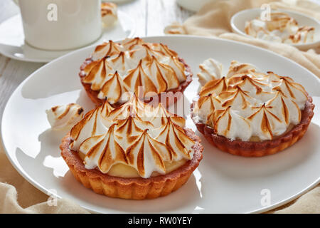 close-up of meringue mini tarts with browned meringue peaks served with tea, mini marshmallows and brown organic sugar on wooden table with light beig Stock Photo