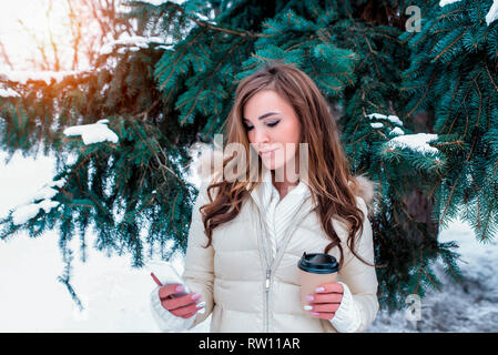 Beautiful girl in winter forest with long hair in her hands holding a mug, a mobile phone reading message online application in social networks Stock Photo