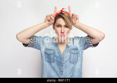 Portrait of funny beautiful young woman in casual blue denim shirt with makeup and red headband standing with horns hands on head and looking at camer Stock Photo