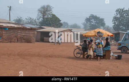 People relaxing under a old and broken sunscreen as a biker is passing by on a small dirty street in rural Ghana. Stock Photo