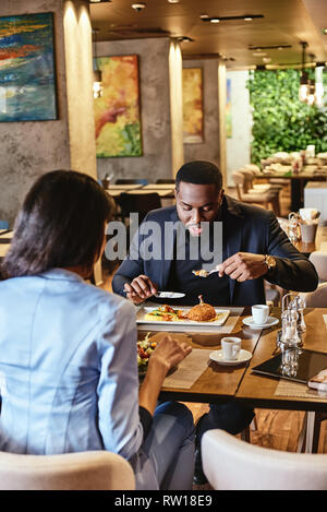 Portrait of co-workers having business lunch at restaurant, sitting at table, discussing project while eating salad and chicken leg. Cozy interior. Ve Stock Photo