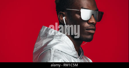 Close up of african man wearing silver outfit and mirrored stylish sunglasses with earphone. Young guy styled in futuristic outfit in studio. Stock Photo