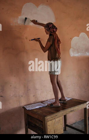 A photo of a woman painting white clouds on a wall of a local elementary school in Ghana, Africa Stock Photo