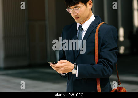 Asian businessman walking outdoors using mobile phone. Korean man walking and using a smart phone outdoor. Stock Photo
