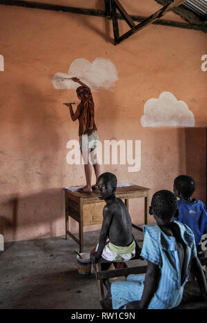 A photo of a woman painting white clouds on a wall of a local elementary school in Ghana, Africa. Students are sitting nearby and watching. Stock Photo