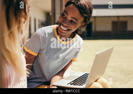 Smiling female students sitting at college campus with laptop computer. Two young students sitting at high school campus with laptop. Stock Photo