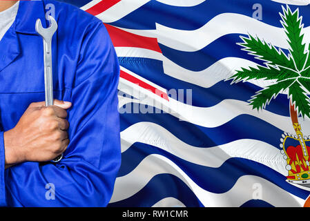 Mechanic in blue uniform is holding wrench against waving British Indian Ocean Territory flag background. Crossed arms technician. Stock Photo