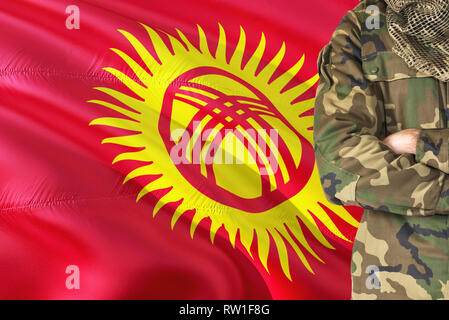 Crossed arms Kyrgyz soldier with national waving flag on background - Kyrgyzstan Military theme. Stock Photo