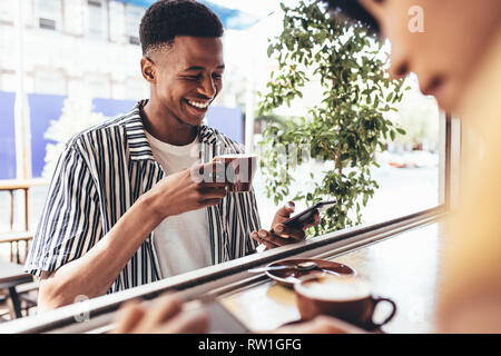 Smiling young african man having coffee and using mobile phone at cafe. Young guy updating his social media status from a cafe. Stock Photo