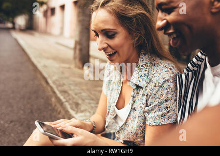 Smiling man and woman using mobile phone sitting outdoors by the street. Friends looking at mobile phone and smiling. Stock Photo