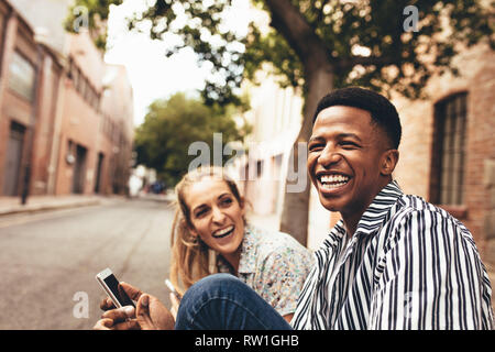 Cheerful multiracial male and female friends sitting outdoors with mobile phone. Happy young friends outdoors. Stock Photo