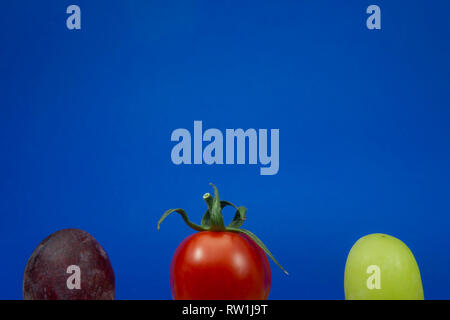 One cherry tomato red and green grapes on a blue background viewed in close-up Stock Photo