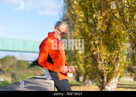 Side view of a senior runner man leaning on fence while testing exercise in a mobile phone outdoors in a sunny day Stock Photo