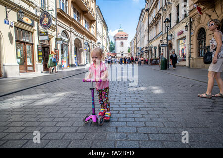 Little girl rides on scooter along the cobblestone streets of Main Market Square in Krak—w Old Town with Brama Florianska, city wall gate, behind them Stock Photo