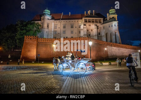 An illuminated horse drawn carriage carries passengers along cobblestone road with Wawel Royal Castle looming above them,  Krak—w, Lesser Poland Voivo Stock Photo