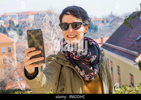 Portrait of a beautiful young woman in warm clothes takes selfie on a smartphone in the city street background, half body. Stock Photo