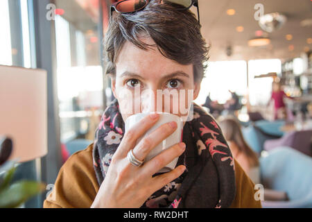 Portrait of a beautiful young woman in warm clothes sitting in cafe and drinkg coffee, blurred background, head and shoulders Stock Photo