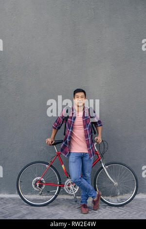 Young man standing with his bike against a gray wall Stock Photo