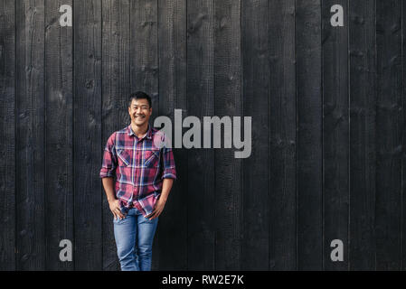 Smiling young Asian man leaning against a dark wall outside Stock Photo
