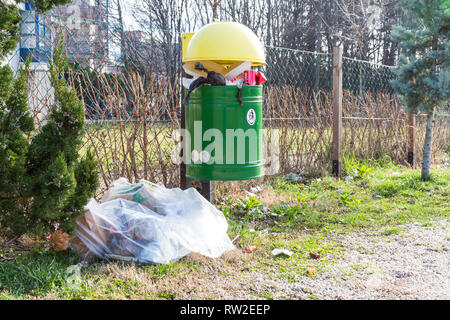 Trash bin full of garbage with a bag of rubbish next to it at playground Stock Photo