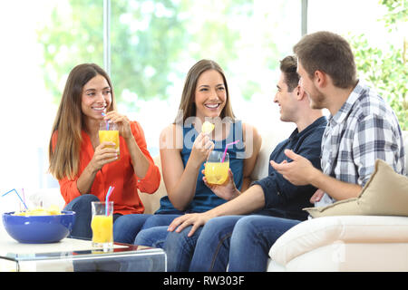 Group of happy friends talking eating chips and drinking refreshments sitting on a couch in the living room at home Stock Photo