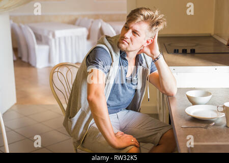 young cute sad man sitting at a table with a plate leaning on his arm against kitchen Stock Photo