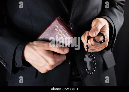 Hands of a christian priest dressed in black holding a crucifix and New Testament book. Religious person with Bible and prayer beads, low-key image. Stock Photo