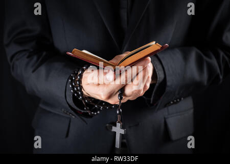 Hands of a christian priest dressed in black holding a crucifix and reading New Testament book. Religious person studies Bible and holds prayer beads, Stock Photo