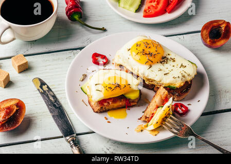 Sandwiches with vegetables and fried egg on white plate, cup of coffee and some fruits over wooden background. Stock Photo