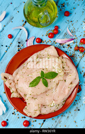 high angle view of some raw marinated chicken fillets in a brown earthenware plate, placed on a blue rustic wooden table next to a glass cruet with ol Stock Photo