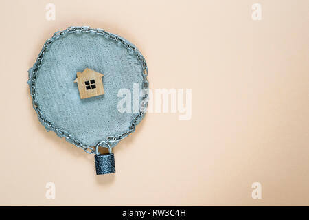 Imitation of a house made of wood in a chain on a lock on a gray piece of concrete on a beige pastel background. The concept of protection of property Stock Photo