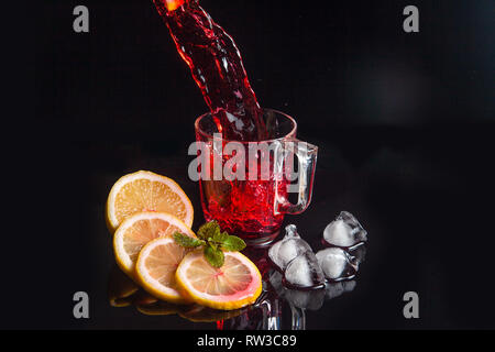 Summer iced drink - hibiscus cold tea with ice, lemon and mint. Hibiscus cold tea pouring into glass with ice isolated on reflective black surface. Hi Stock Photo
