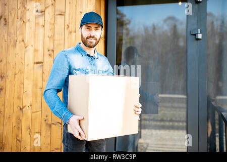 Portrait of a delivery man in blue workwear standing with packaging near the modern house Stock Photo