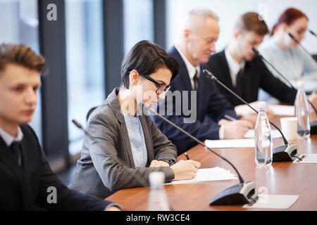 Side view portrait of young businesswoman taking notes while sitting at table in conference room during business seminar, copy space Stock Photo