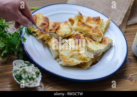 Traditional turkish borek in a enamel dish on wooden table.Woman's hand  taking borek with fork. Stock Photo