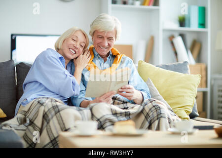 Portrait of loving mature couple reading book together sitting on sofa at home, copy space Stock Photo