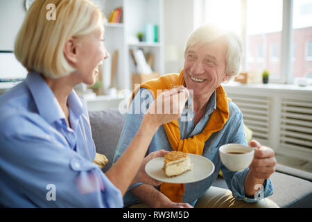 Portrait of happy mature couple enjoying tea and cake in sunlit apartment, copy space Stock Photo