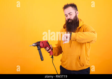 Bearded man wearing yellow hoodie hoding power drill and looking at his watch over yellow background. Handsome man. Worker. Stock Photo