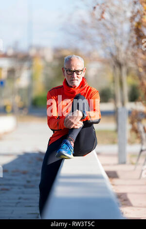 Front view of a senior man wearing sportswear and eyeglasses sitting on a fence relaxing while looking camera outdoors in a sunny day Stock Photo