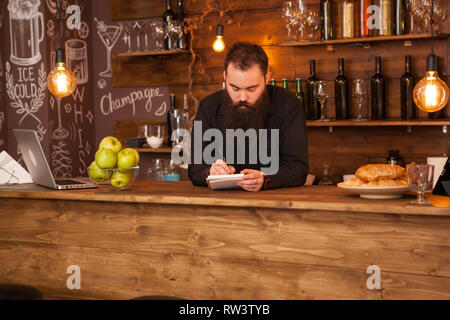 Bearded hipster bartender taking notes behind bar counter Stock Photo