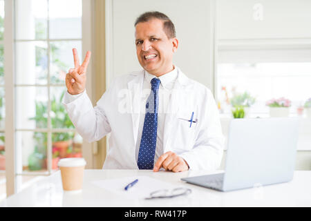 Middle age doctor man wearing white medical coat working with laptop at the clinic smiling with happy face winking at the camera doing victory sign. N Stock Photo