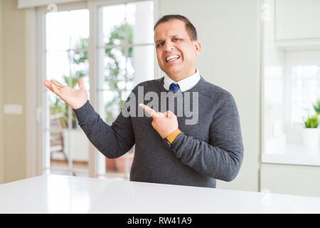 Middle age man sitting at home amazed and smiling to the camera while presenting with hand and pointing with finger. Stock Photo