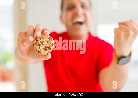 Middle age man eating chocolate chips cookies at home screaming proud and celebrating victory and success very excited, cheering emotion Stock Photo