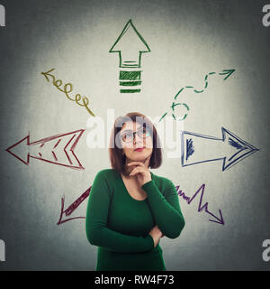 Ingenious, pensive woman holding hand under chin thinking to choose a direction as colored arrows on the gray wall are pointed to different directions Stock Photo