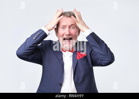 Oh no concept. Surprised, shocked man in blue suit holding hands up near head isolated on grey background Stock Photo