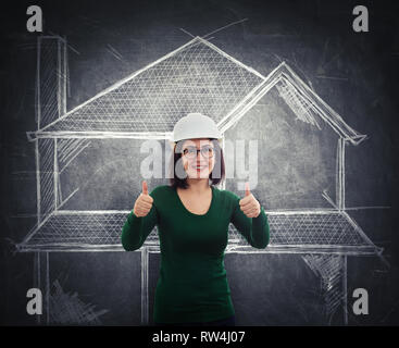 Happy cheerful young woman engineer wearing protective helmet and glasses smiling showing thumbs up gesture looking to camera standing over blackboard Stock Photo
