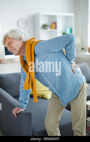 Portrait of contemporary senior man suffering from back pain leaning on sofa Stock Photo