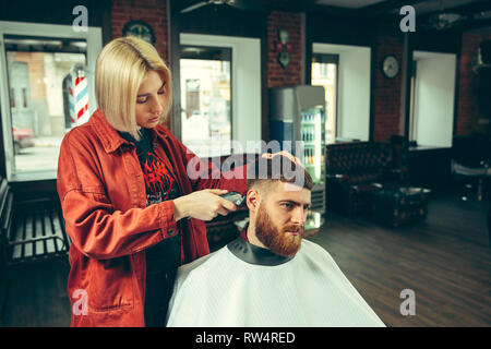 Client during beard shaving in barbershop. Female barber at salon. Gender equality. Woman in the male profession. Stock Photo